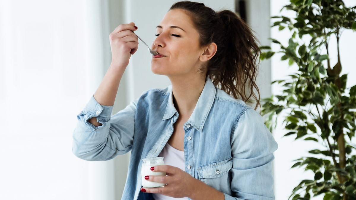 A woman with her eyes closed takes a bite of plain Greek yogurt from a jar she's holding.