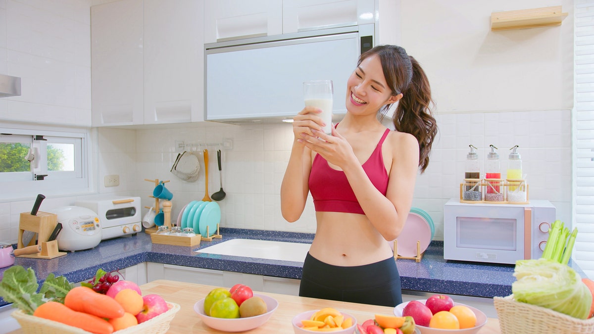 A woman in an exercise outfit holds a glass of milk in both hands while standing in a kitchen with fruits and vegetables.