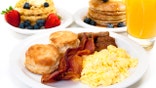 A breakfast plate with scrambled eggs, bacon and buttermilk biscuits. Waffles, pancakes and orange juice are in the background.