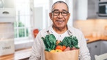 A man wearing glasses holds a grocery bag with fruits and vegetables while standing in a kitchen.
