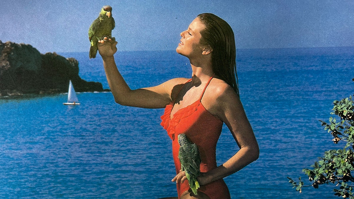 Christie Brinkley wearing a red swimsuit holding parrots by the ocean.