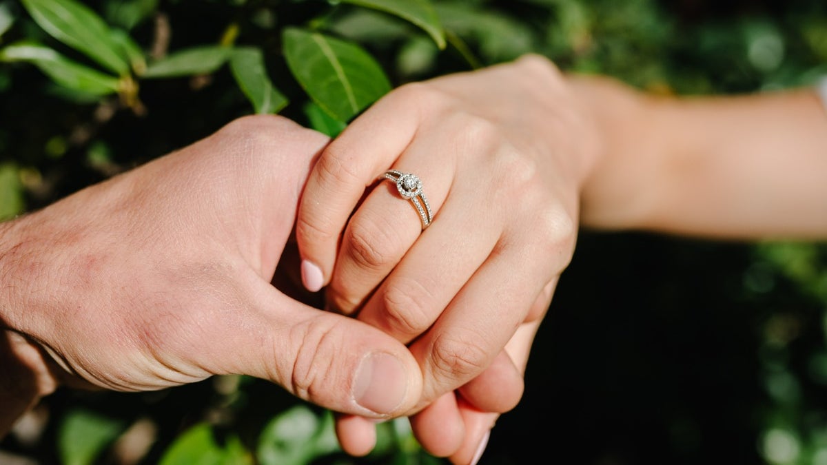 Two hands with one wearing an engagement ring in front of a plant