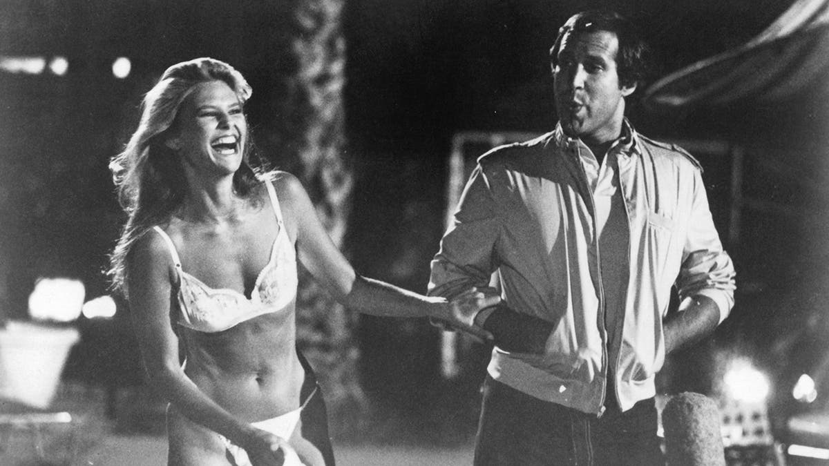 Christie Brinkley laughing in a white bikini walking next to Chevy Chase.