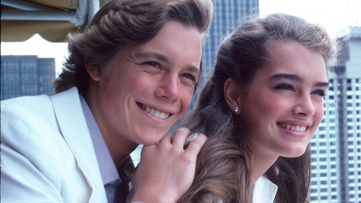 Christopher Atkins smiling and holding onto Brooke Shields as they both wear white.