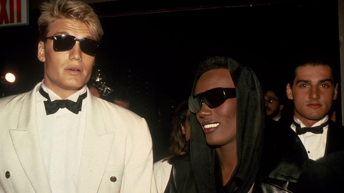 Dolph Lundgren in a white suit with sunglasses and a bowtie standing next to a smiling Grace Jones.