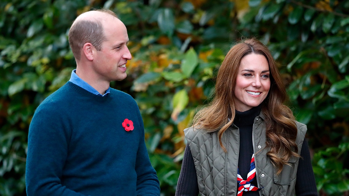 Prince William wearing a blue sweat and a red floral pin next to Kate Middleton in a hunter green puffy vest.