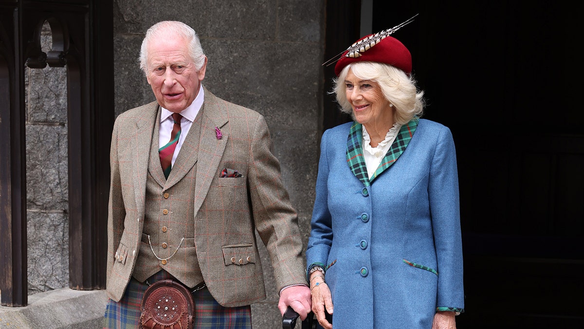 King Charles wearing a Scottish kilt and a brown blazer walking next to Queen Camilla in a bright blue coat dress.