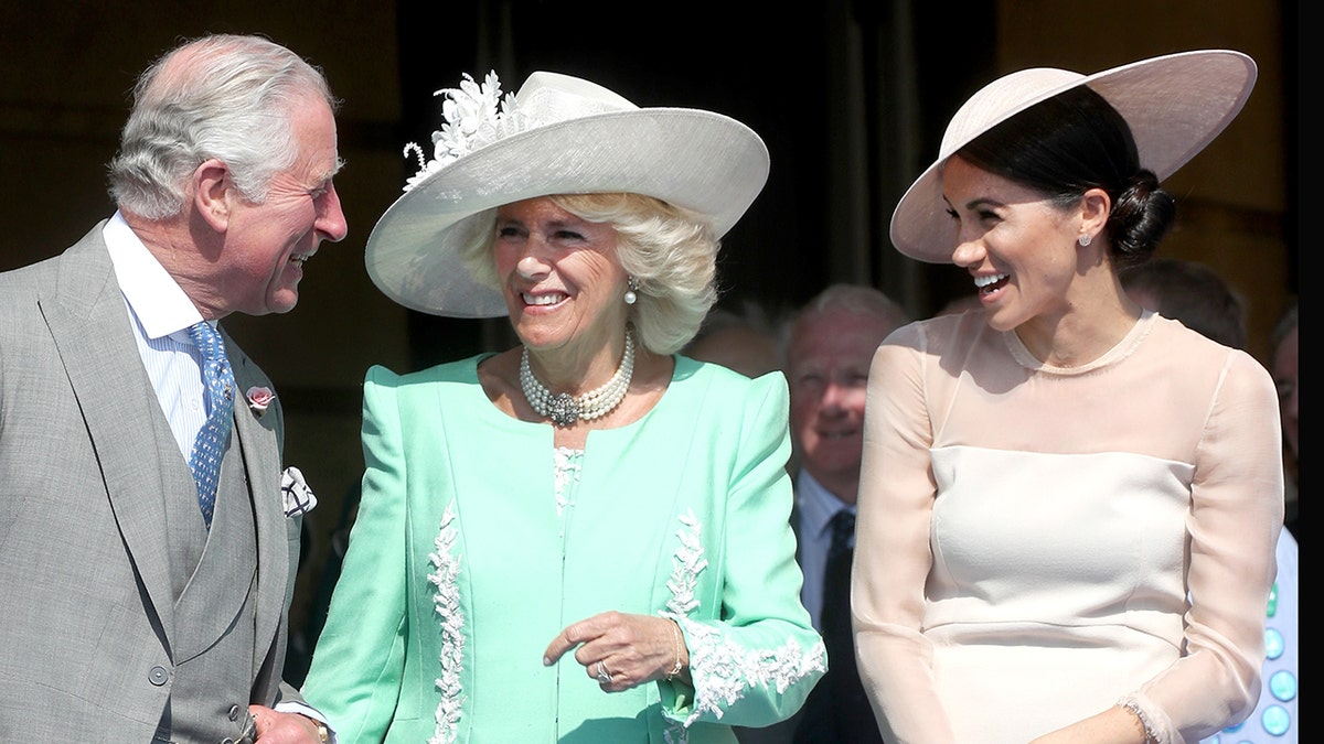 King Charles, Queen Camilla and Meghan Markle laughing in formal wear outside.