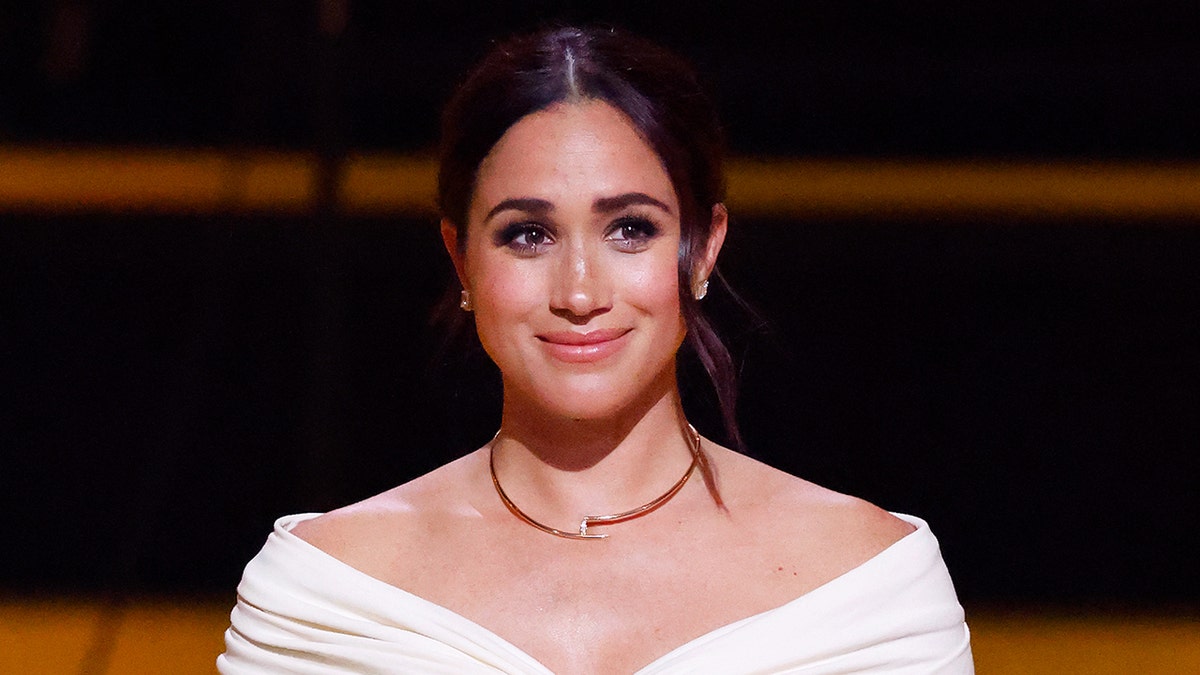 Meghan Markle sourit vêtu d'un haut blanc et d'un collier en or.