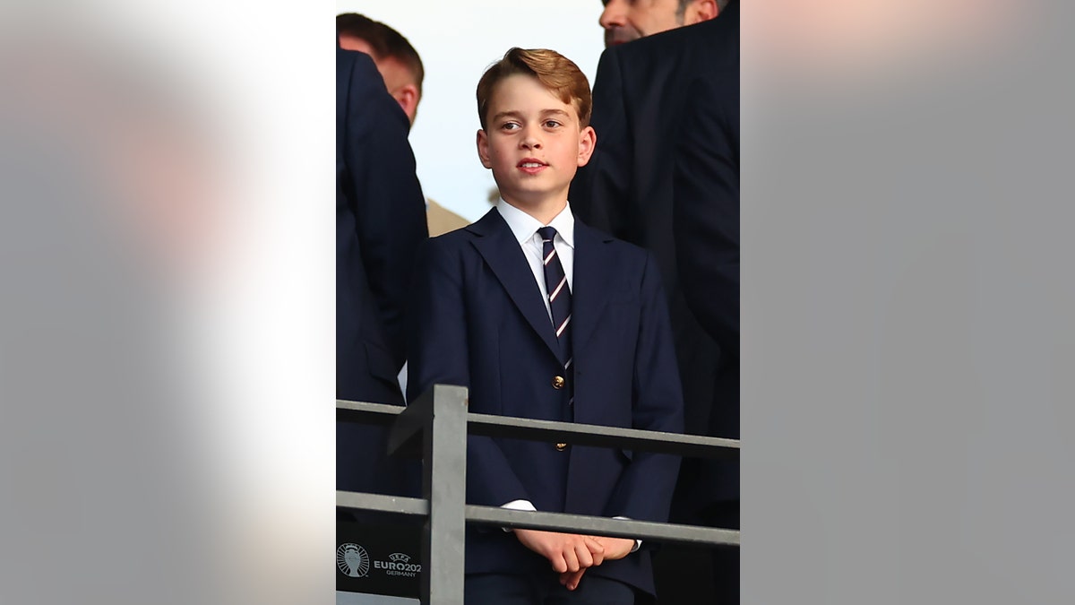 Prince George in a blue suit and striped tie looking up from a crowd.