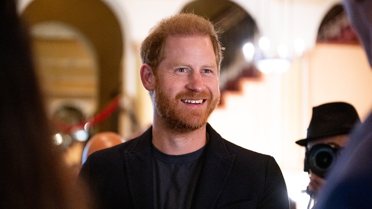 A close-up of Prince Harry smiling wearing a black blazer and a matching shirt speaking to guests.