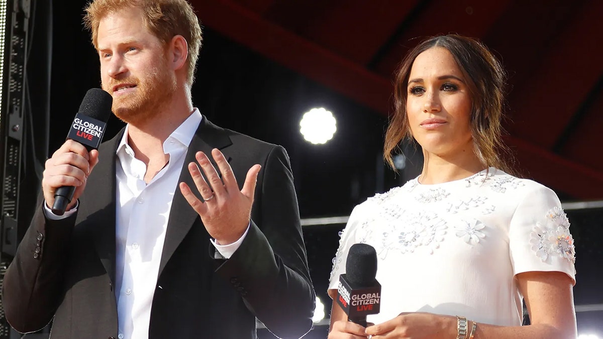 Prince Harry talks to the microphone next to Meghan Markle, wearing a dark suit and a white shirt, in a white shirt, wearing a white dress, holding the microphone and looking at him.