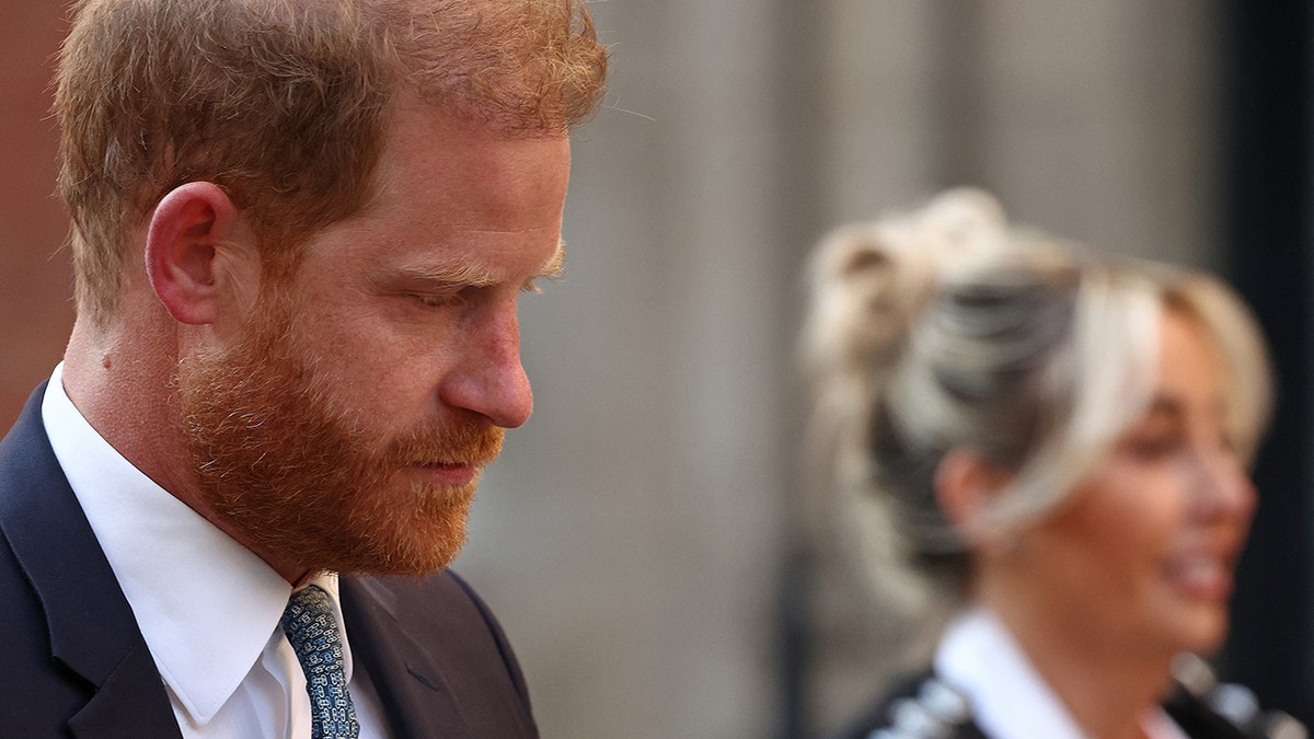A close-up of Prince Harry looks gloomy next to a smiling blonde woman.