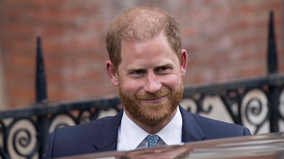 A close-up of Prince Harry smiling in a suit in front of the car.