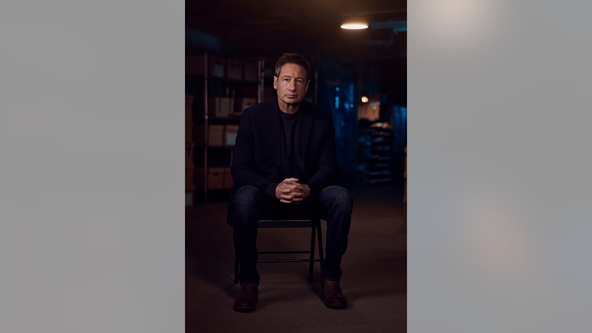 David Duchovny sitting down in a dark suit and matching blazer and shirt looking serious.