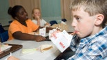 A boy drinks milk through a straw at school while two girls sit at his table with their milks and lunch.