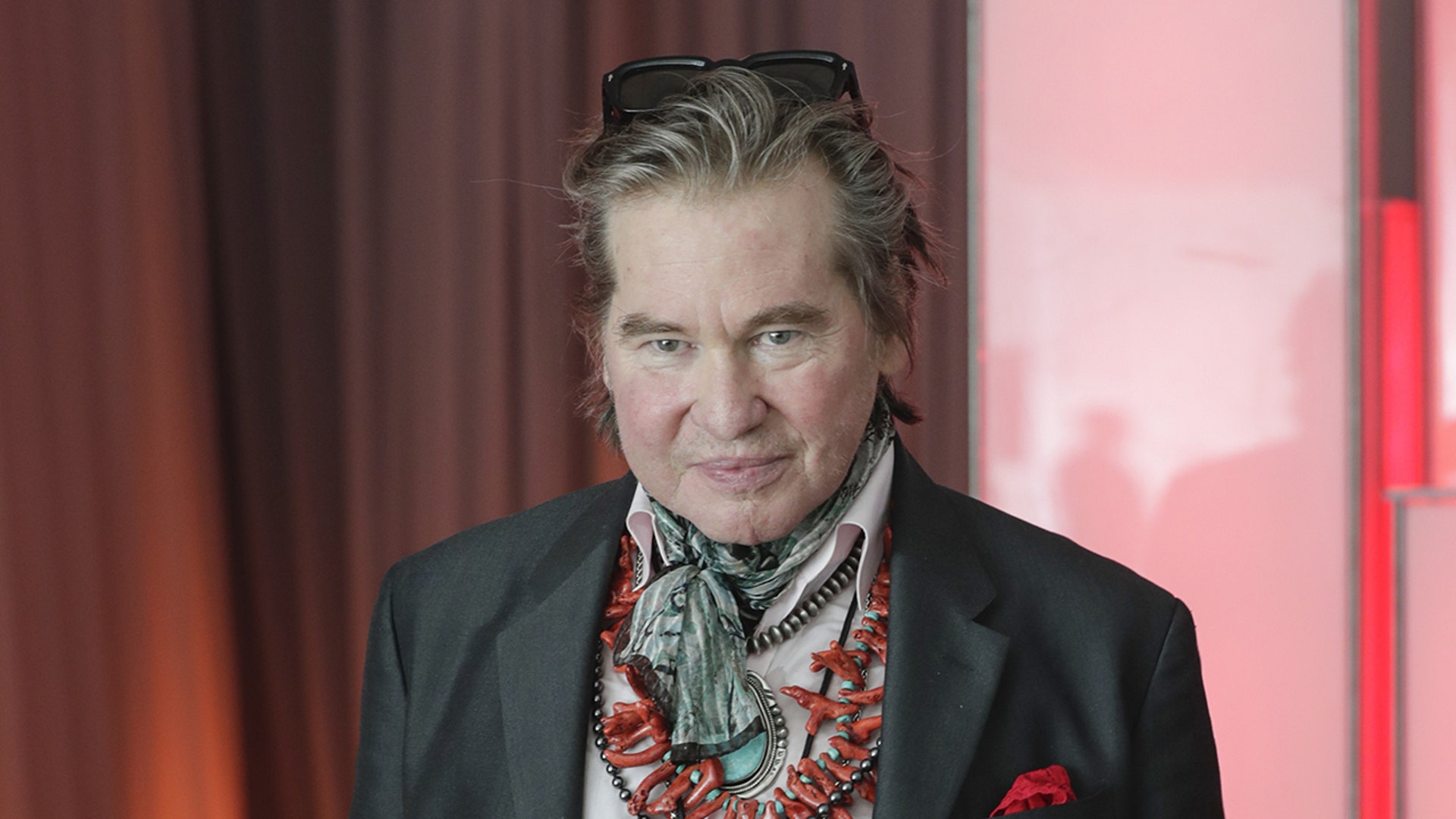 Actor Val Kilmer wears grey blazer and pink shirt.