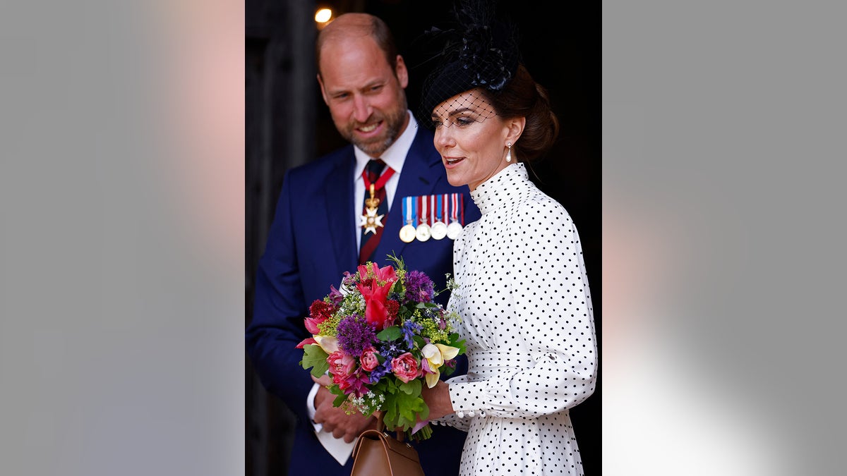 Prince William in a navy suit looking at Kate Middleton holding a bouquet of flowers wearing a polka dot dress and a black fascinator.
