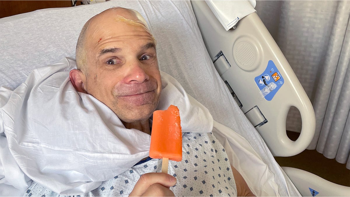 Rand Laycock sits upward in a hopsital bed wearing a gown, smiling into the camera with eyes wide as he holds an orange popsicle thay has been bitten into