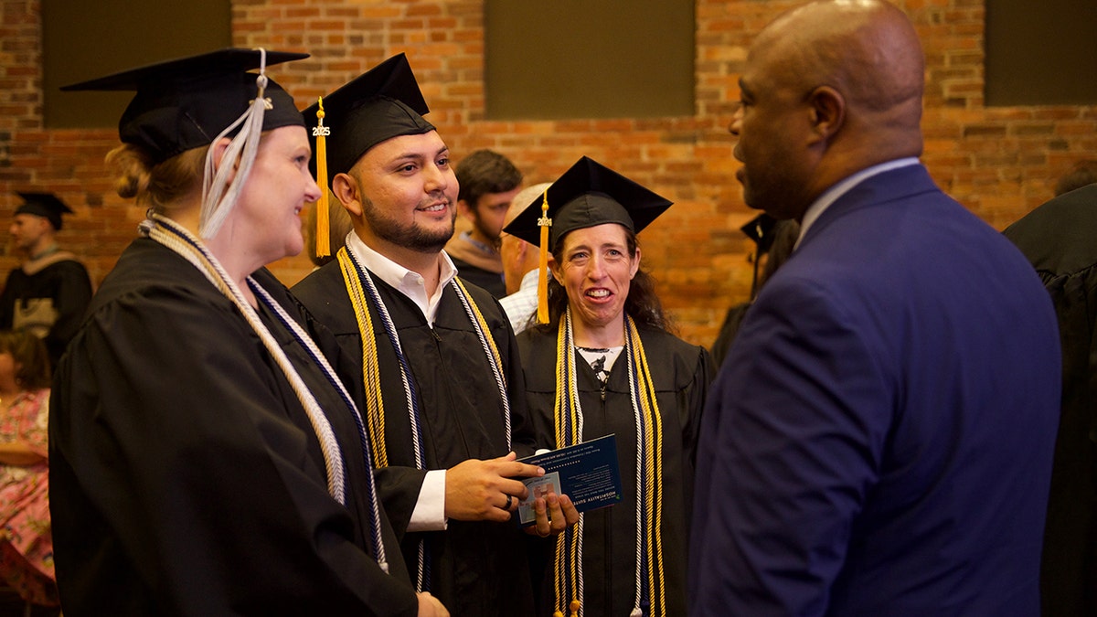Three university graduates with their caps and dresses talk to Andre 'Kennebrew, which wears a suit.