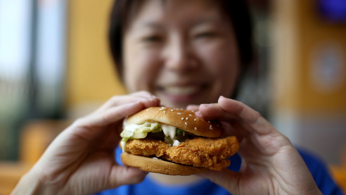 An Asian woman, whose face is out of focus, holds a chicken sandwich.