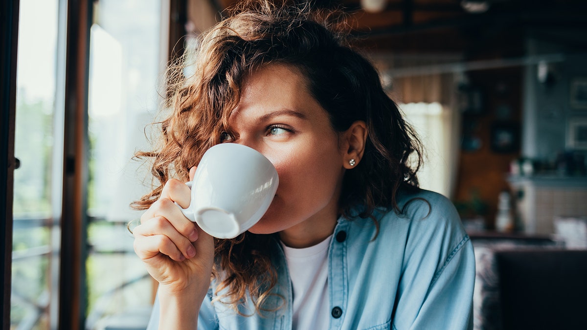 Portrait of happy beautiful young woman wearing blue shirt drinking coffee from a white cup sitting at table in restaurant looking out the window on summer day.