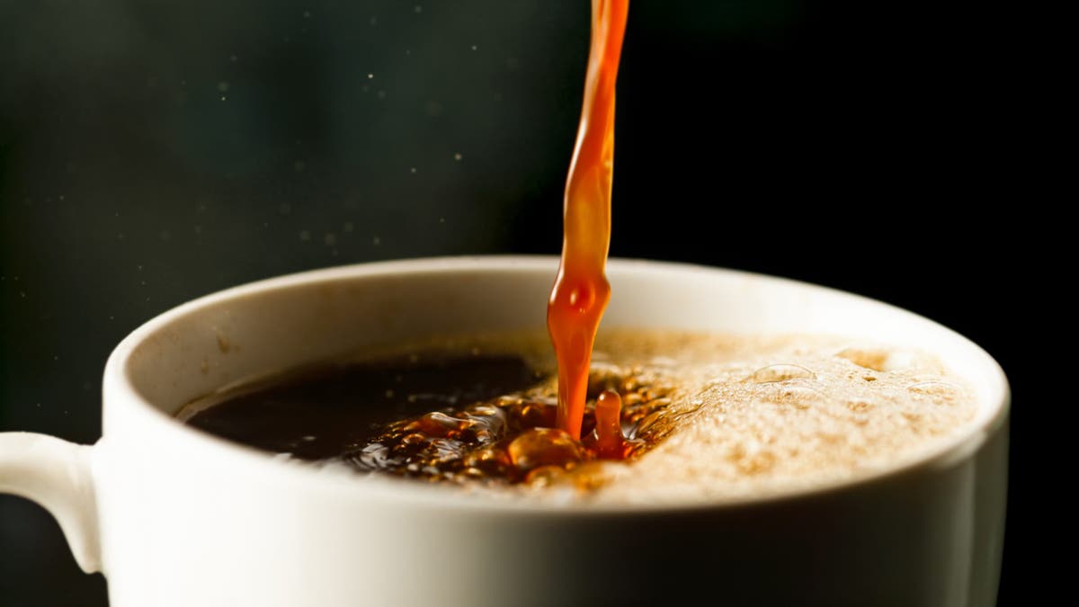 Coffee being poured into a white cup from above from a golden brown stream as foam sits on the right side of the cup