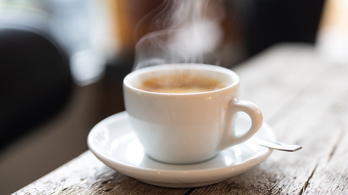 Steam rising from a white cup of hot coffee with a spoon on a saucer over a wooden table in the cafe. Close-up of a refreshing hot cup of a coffee at a cafe.