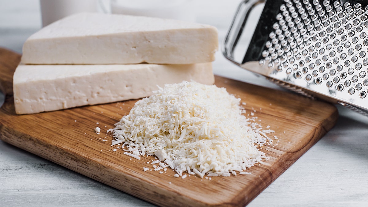 Cotija cheese, in blocks and shredded, is shown on a cutting board next to a cheese grater.