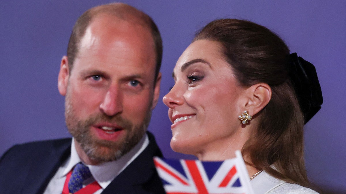A close-up of Prince William telling something to a smiling Kate Middleton with a Union Jack flag in front of her.