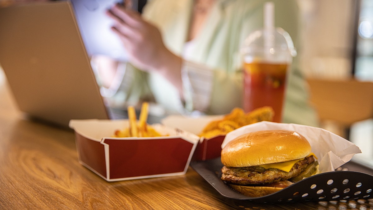Businesswoman working at laptop with fast food on restaurant table. Pictured are fires, chicken tenders, a beverage with a straw, and a hamburger