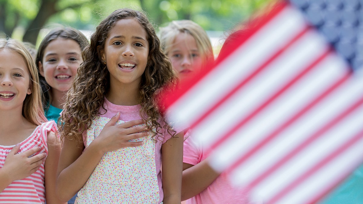 Biracial African American and Caucasian elementary age little girl is standing outdoors with diverse classmates. Students are reciting Pledge of Allegiance to the American Flag. For Flag Day use