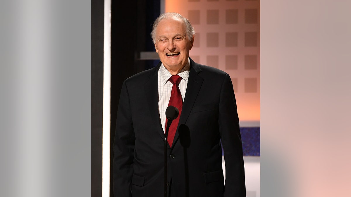 Alan Alda wearing a black blazer and a red tie on stage smiling.