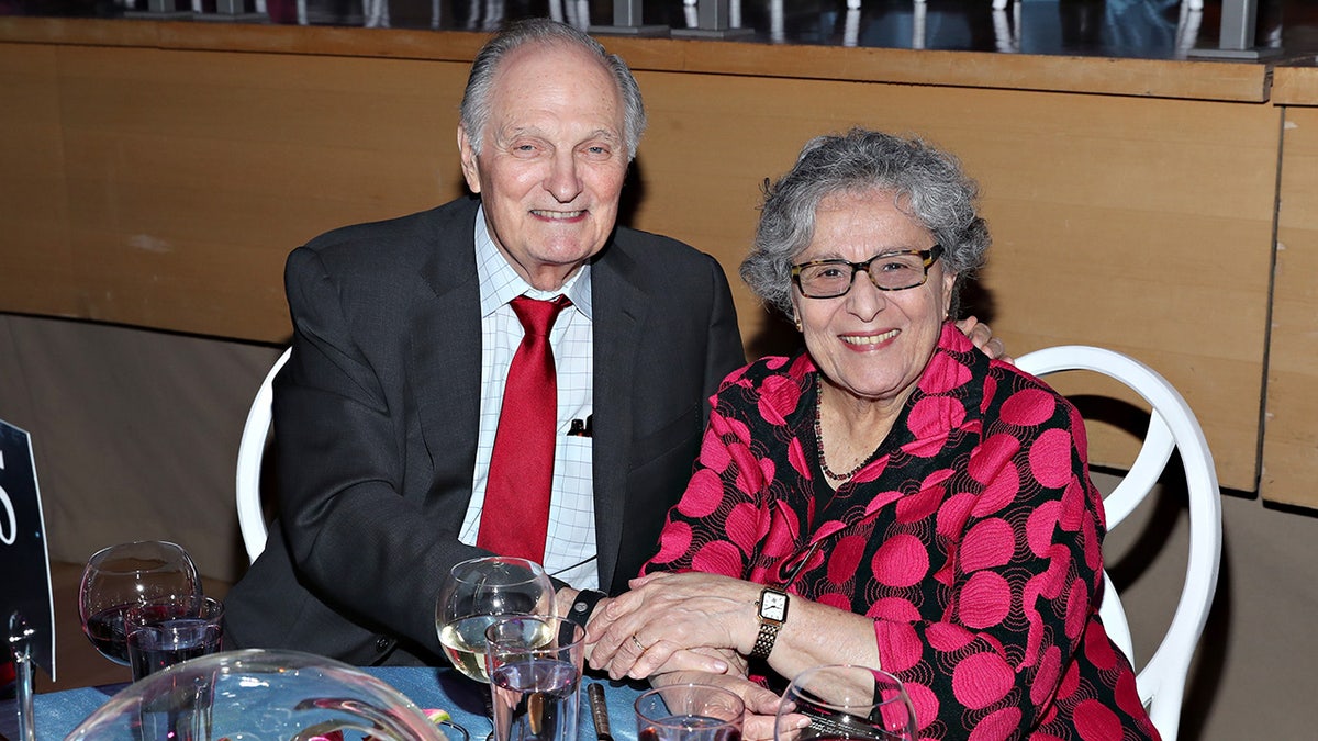 Alan Alda in a suit and red tie sitting next to his wife at a dining table.