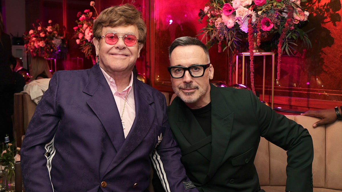 Elton John and his husband sitting together and smiling inside a venue with florals.