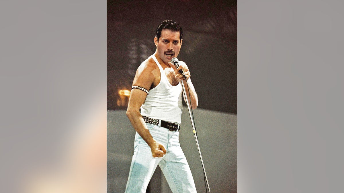 Freddie Mercury singing on stage wearing a white tank top and matching white jeans.