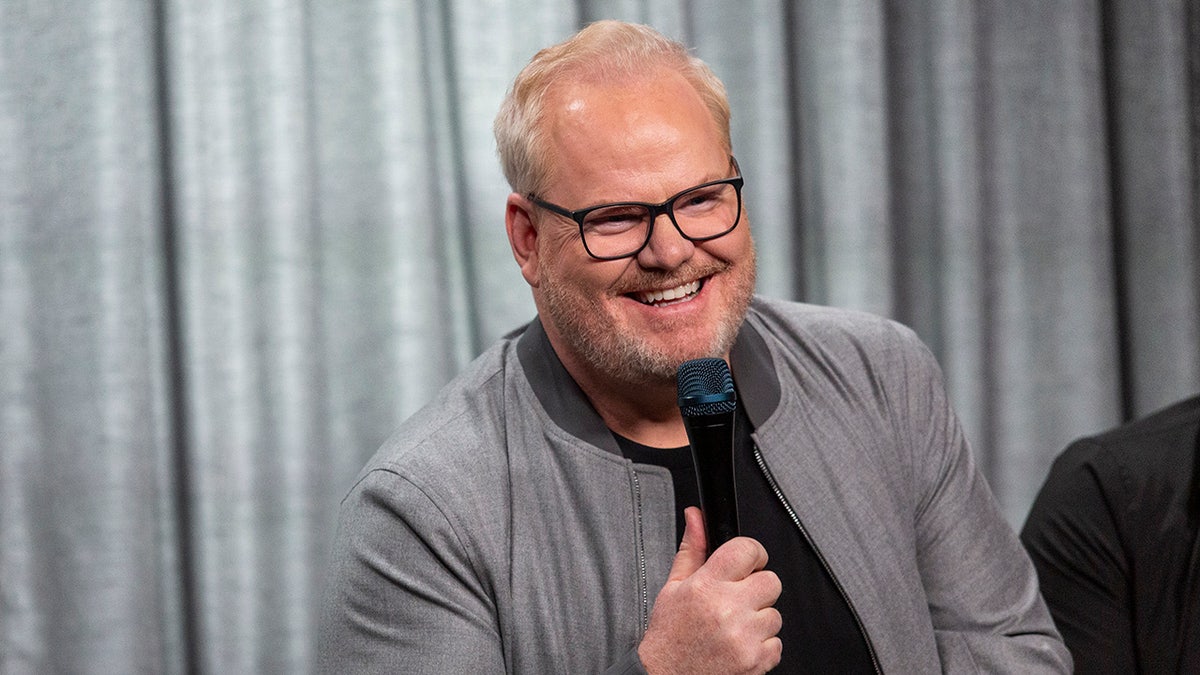 A close-up of Jim Gaffigan wearing a grey sweater with a black shirt smiling and holding a mic.