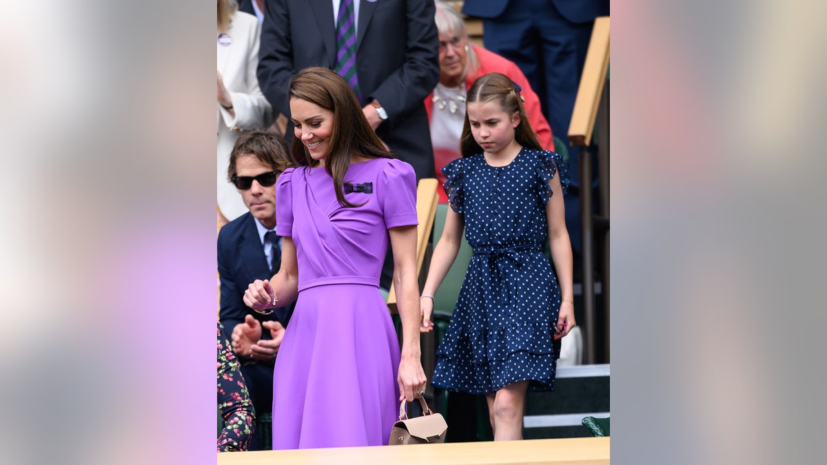 Kate Middleton wearing a bright purple dress walking in front of Princess Charlotte who is wearing a polka dot dress.