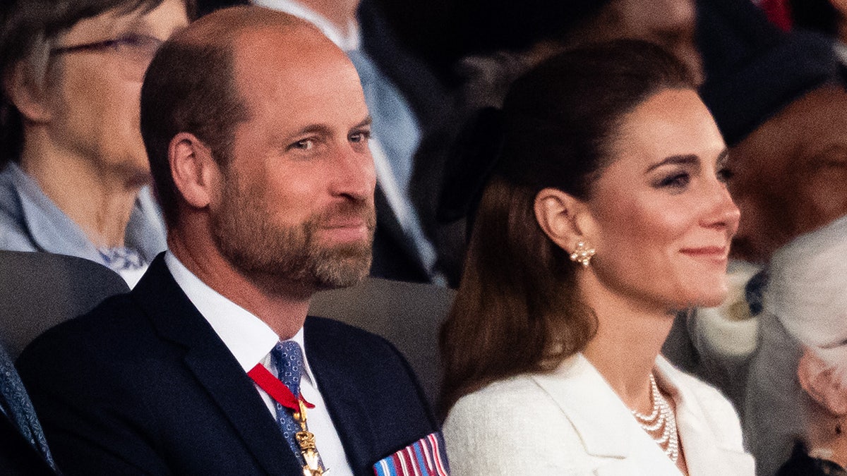 Prince William smiling in a suit with medals sitting next to Kate Middleton wearing a white coat dress and pearls.