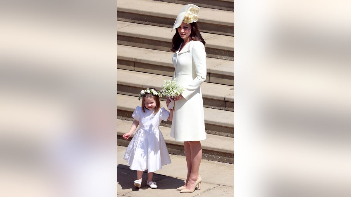 Kate Middleton wearing a white coat dress holding onto Princess Charlotte's hand. Kate's daughter is wearing a white dress with a flower crown.