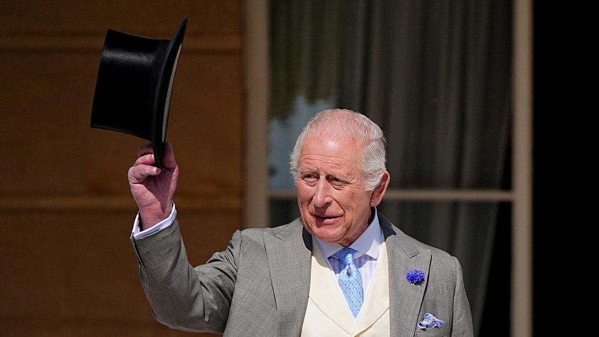 King Charles waving his top hat while wearing a grey and beige suit.