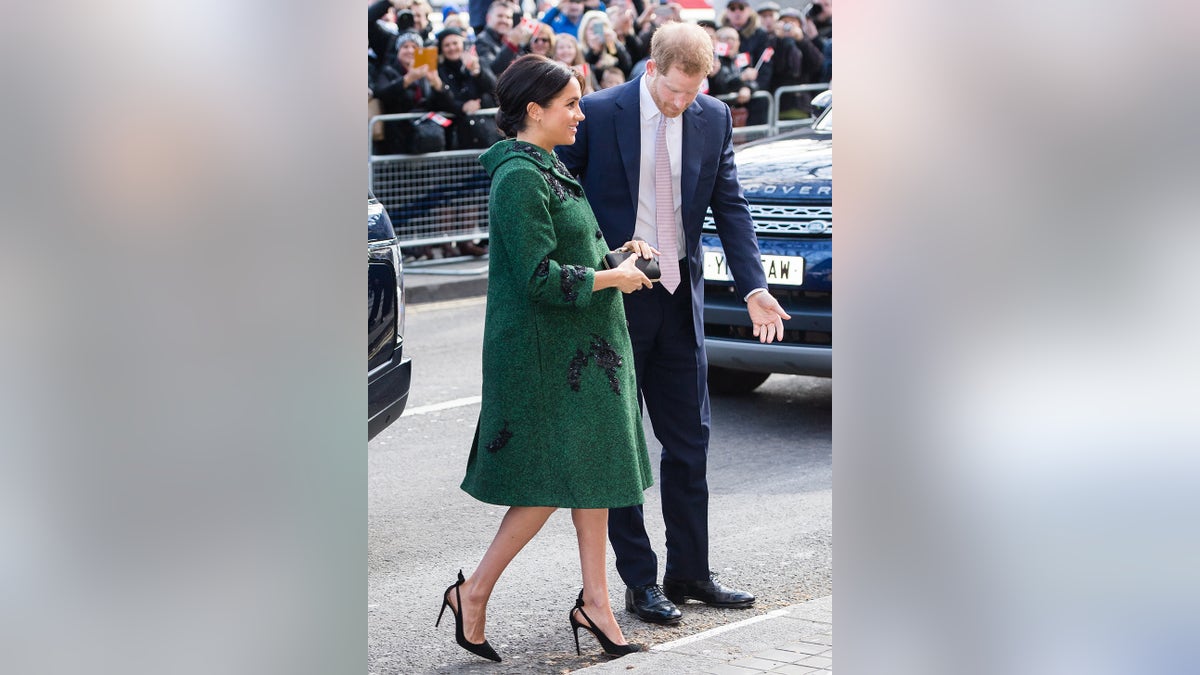 Meghan Markle wearing a green coatdress and high heels while pregnant holding on to Prince Harry in a dark blue suit and tie.