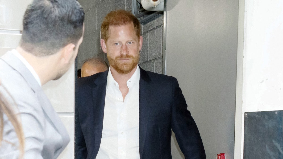 A close-up of Prince Harry walking forward as a security guard looks on.
