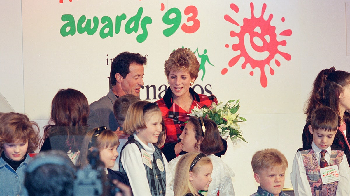 Sylvester Stallone and Princess Diana posing together behind a group of children holding flowers.