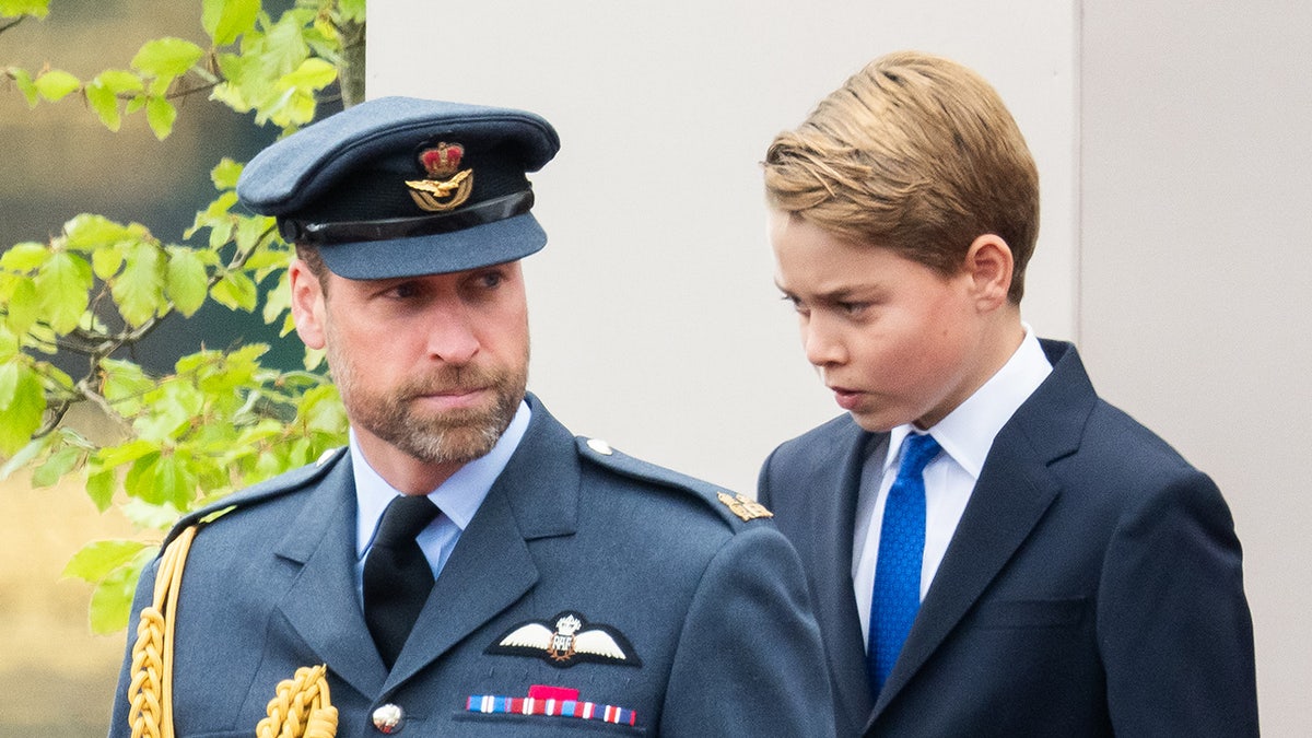 Prince William in uniform walking in front of Prince George in a suit.