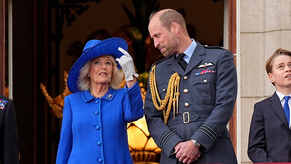 Prince William smiling and chatting with Queen Camilla on the palace balcony who is looking up.