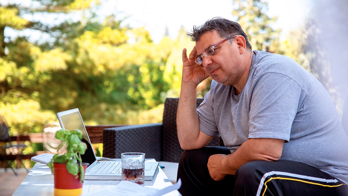An overweight man sits outside on a chair, his hand pressed to his head. He looks disgruntled.