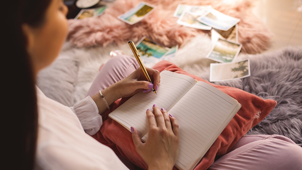 A young woman sits on her bed, writing in her journal with her left hand.