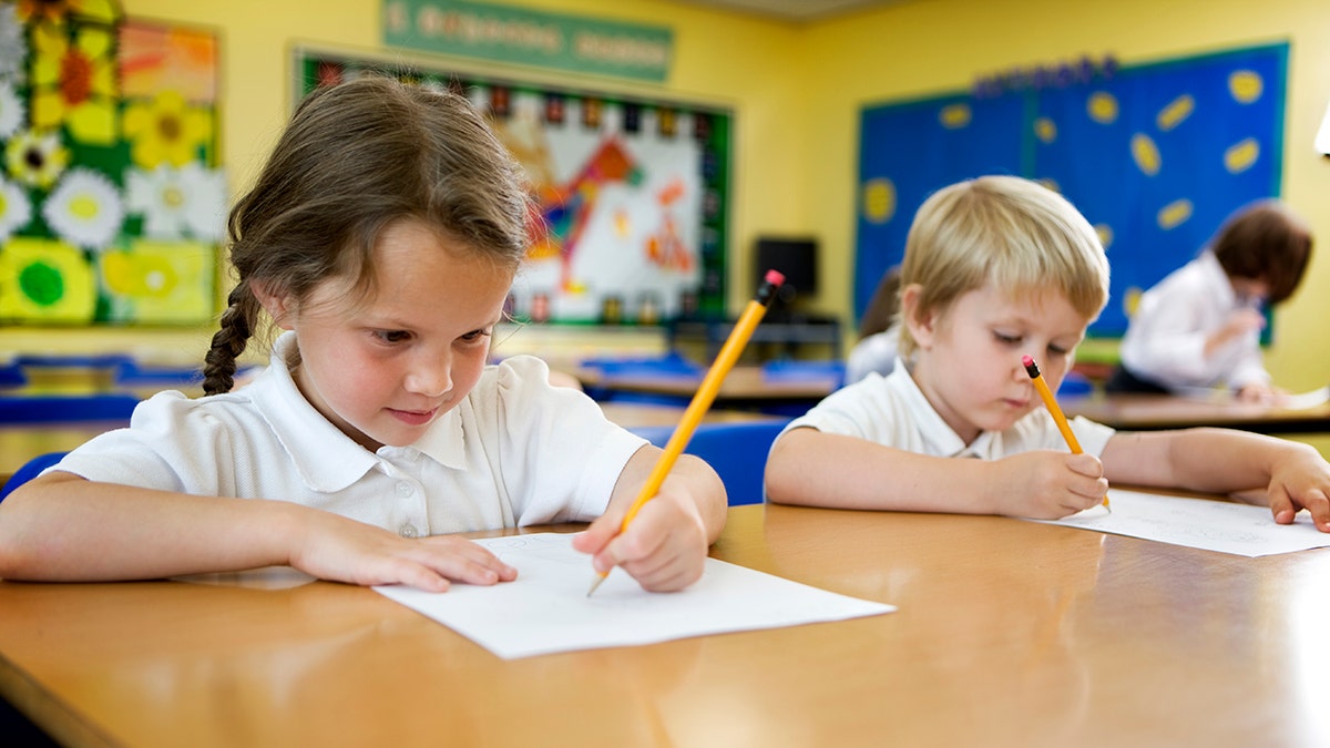 A little girl and little boy write on paper in their classroom at their desks. The little girl is writing with her left hand.