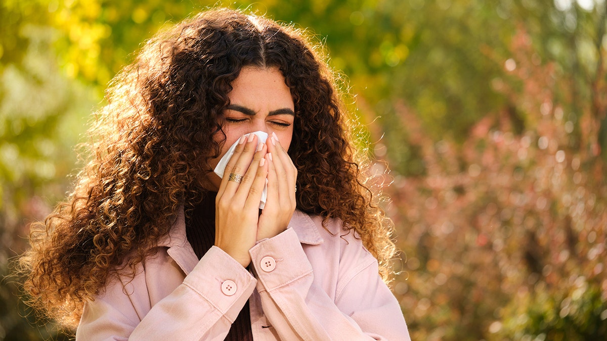 A woman with brown curly curly holds a tissue to her nose with both hands, covering the bottom half of her face. Her eyes are closed as she blows her nose and is positioned in the foreground of a green backdrop set in nature